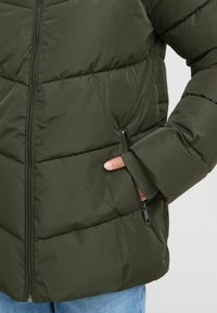 Temno zelena puffer jakna s prešitejšimi deli, ki ima sprednji zadrgo in stransko žep. Material deluje vodoodporen z gladko teksturo.