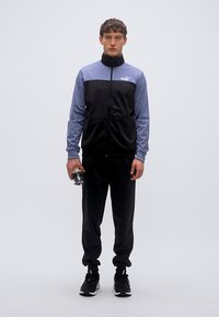 Μαύρη και γαλάζια φόρμα με φερμουάρ στο μπουφάν, που διαθέτει ψηλό γιακά και ριμπ μανσέτες, συνδυασμένη με μαύρο παντελόνι και αθλητικά παπούτσια.