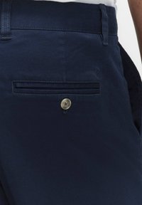 Marineblaue Hose mit glatt strukturiertem Material, ausgestattet mit einer Gesäßtasche mit Knopf und dezenten horizontalen Falten oberhalb der Tasche.