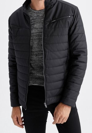 Homme portant une veste matelassée noire par-dessus un pull en maille gris avec un pantalon noir, debout devant un fond blanc uni.