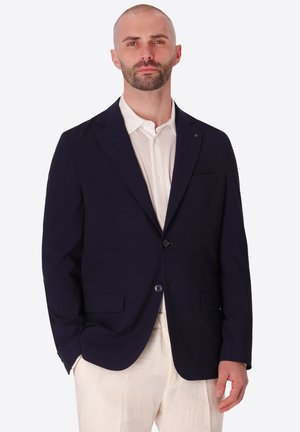Uomo che indossa un blazer blu scuro sopra una camicia bianca e pantaloni beige, in piedi con una mano in tasca contro uno sfondo neutro.