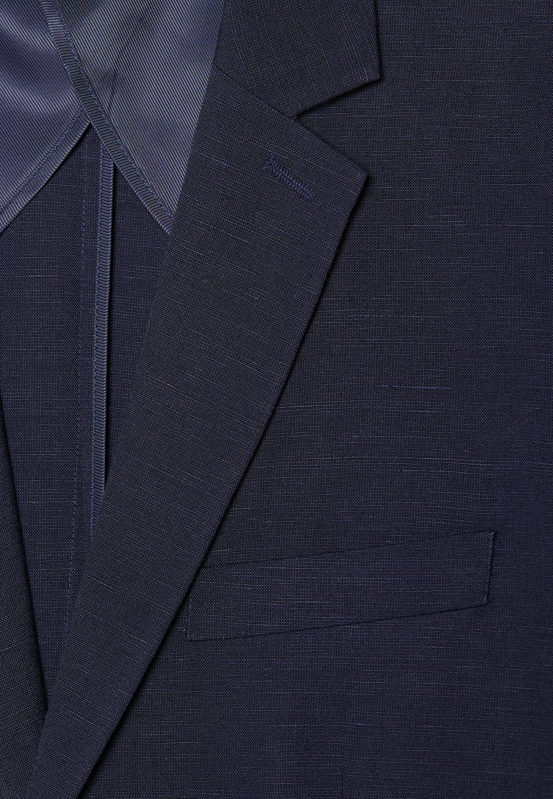 Veste de costume bleu marine en tissu texturé, avec des revers à cran et une poche poitrine unique. Les accents comprennent une doublure intérieure contrastante et des coutures.