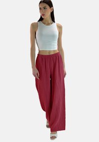 Wijdvallende, getextureerde rode broek met een elastische tailleband, gecombineerd met een witte, aansluitende tanktop. Het model draagt witte sandalen.