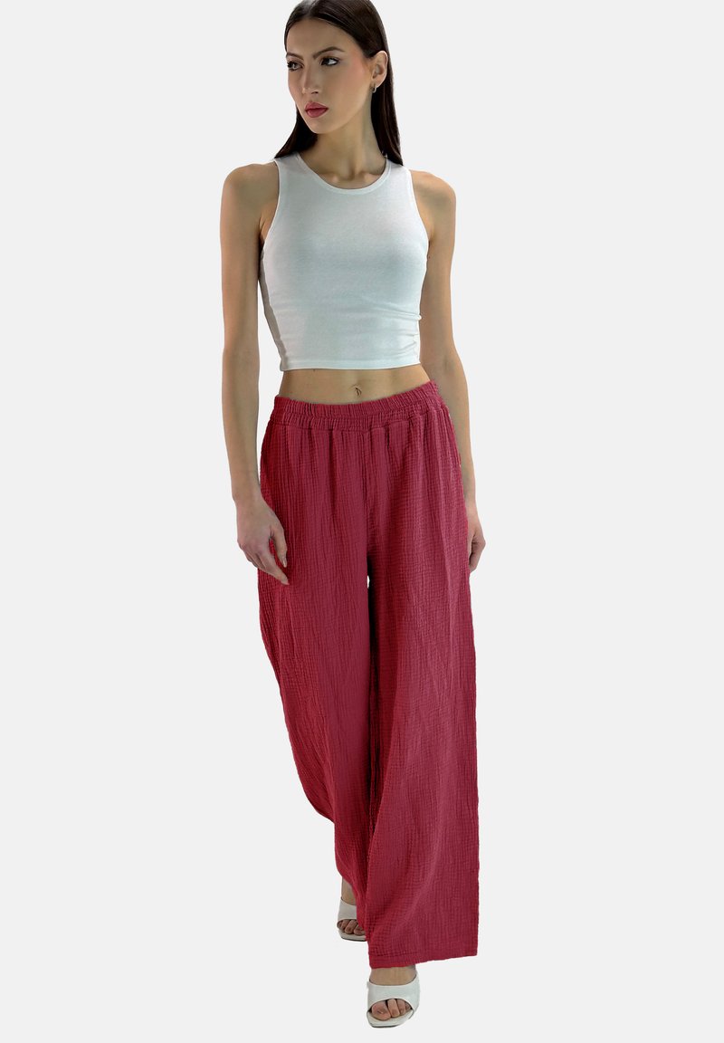 Wijdvallende, getextureerde rode broek met een elastische tailleband, gecombineerd met een witte, aansluitende tanktop. Het model draagt witte sandalen.