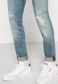 Blå slitna jeans med fransade hål, uppvikta fållar, tillsammans med vita sneakers med röda detaljer.