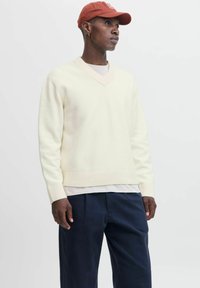 Man wearing a cream V-neck sweater, navy pants, beige shirt underneath, rust-colored cap, and small hoop earring standing against a white background.