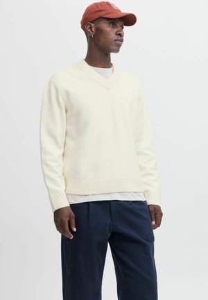 Man wearing a cream V-neck sweater, navy pants, beige shirt underneath, rust-colored cap, and small hoop earring standing against a white background.