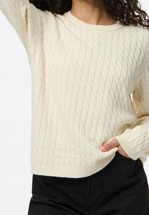 Femme portant un pull crème à torsades avec poignets et ourlet côtelés, associé à un pantalon noir, la main posée dans une poche.