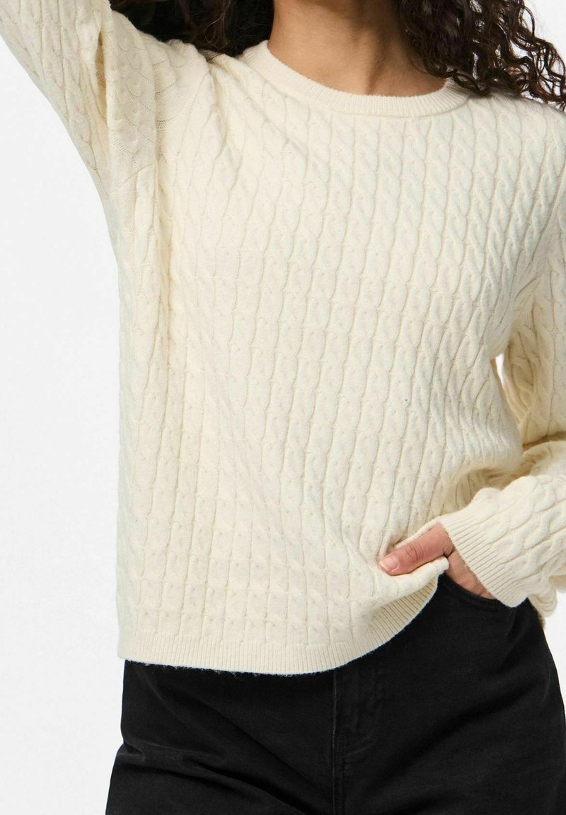 Femme portant un pull crème à torsades avec poignets et ourlet côtelés, associé à un pantalon noir, la main posée dans une poche.