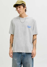 Mlad moški nosi olivno zeleni klobuk, sivo majico s plavim napisom "Originals Studio", srebrno verigo, modre kavbojke in vidne tetovaže na obeh rokah.