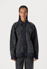Chaqueta de denim en azul oscuro con un patrón texturizado de puntos blancos. Cuenta con un gran cuello, bolsillos frontales y cierre de botones.