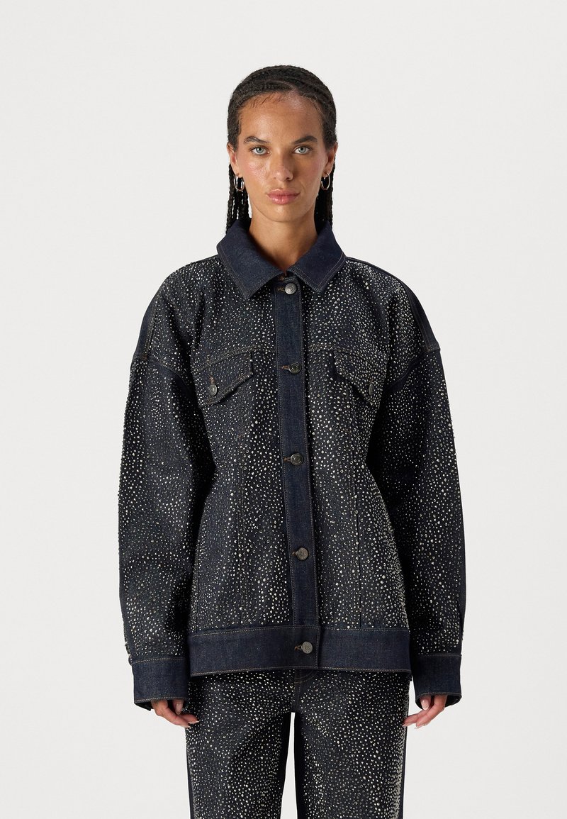 Chaqueta de denim en azul oscuro con un patrón texturizado de puntos blancos. Cuenta con un gran cuello, bolsillos frontales y cierre de botones.