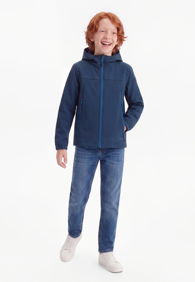 Garçon roux souriant, portant une veste à capuche bleu marine, un jean bleu et des baskets blanches, debout devant un fond blanc.