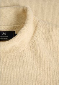 Cremfarbener Strickpullover mit hohem Rippenkragen, der ein strukturiertes Muster und ein kleines Logolabel in der Nähe des Halsausschnitts aufweist.