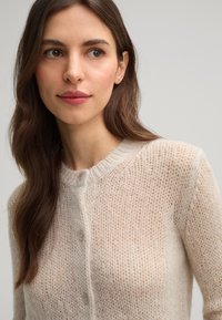Strickjacke in hellbeige, mit Rundhalsausschnitt, Knopfverschluss und einem lockeren, strukturierten Stoff, der eine zarte, luftige Erscheinung hat.