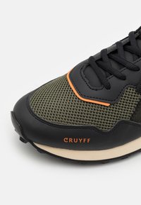 Sneaker pour homme avec un mesh vert olive, une superposition synthétique noire, des accents orange et une semelle intermédiaire couleur crème pour un bon amorti.