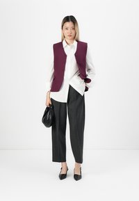 Burgunder vest over hvit skjorte, sammen med skræddersydde grå bukser. Svart håndveske og spisse hæler fullfører antrekket. Enkel design, rene linjer.
