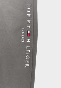 Grauer Stoff mit vertikalem weißen Text "Tommy Hilfiger EST. 1985" und einem kleinen rot-weiß-blauen Tommy Hilfiger-Logo.