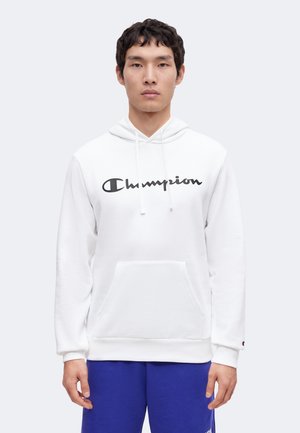 Joven con una sudadera con capucha blanca de Champion con logo negro y pantalones azules, de pie frente a un fondo liso gris claro.