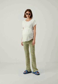 Weiße kurzärmlige Bluse mit einem Knoten an der Taille, grüne gemusterte Schlaghosen und blaue Rutsch-Sandalen, getragen von einem schwangeren Modell.