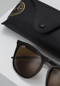 Ochelari de soare negri Ray-Ban cu un cadru cu efect de broască țestoasă și lentile maronii, însoțiți de un toc protector texturat de culoare neagră.