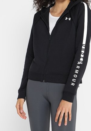 Vrouw draagt een zwarte Under Armour hoodie met ritssluiting, voorzien van een wit logo op de borst en branding op de mouw, gecombineerd met grijze nauwsluitende leggings.