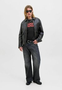 Černá kožená bunda přes černé tričko s červeným textem, v kombinaci s tmavými džíny a černými botami. Model má dlouhé vlasy a sluneční brýle.