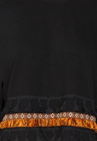Haut noir avec un bord décoratif orné de motifs orange et blancs, agrémenté de glands noirs le long de l'ourlet pour apporter texture et détail.