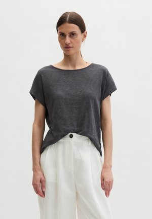 Femme aux cheveux bruns lisses portant un haut ample gris foncé à manches courtes, rentré dans un pantalon blanc taille haute à jambes larges.