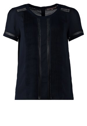 Vrouw met een zwarte blouse met korte mouwen, witte slim-fit broek, taupekleurige enkellaarsjes en ze houdt een tweekleurige handtas met een gouden kettinghengsel vast.