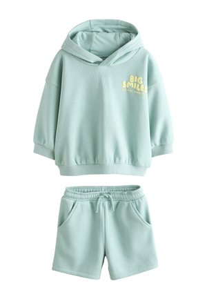 Junges Kind trägt einen hellblauen Hoodie mit Schriftzug draußen auf einer Graslandschaft unter klarem blauem Himmel und schaut nachdenklich in die Ferne.
