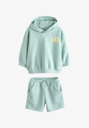 Junges Kind trägt einen hellblauen Hoodie mit Schriftzug draußen auf einer Graslandschaft unter klarem blauem Himmel und schaut nachdenklich in die Ferne.
