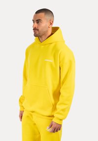Felpa gialla con cappuccio realizzata in morbido misto cotone, caratterizzata da una tasca frontale e logo ricamato sul petto. Texture liscia, vestibilità rilassata.