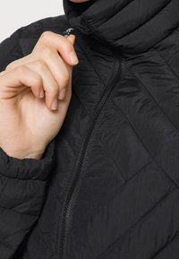 Svart kviltad jacka med hög krage och dragkedja. Har en slät, mjuk yta och en åtsittande design. Ett finger som greppar dragkedjans veck.