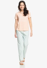 Camiseta de melocotón claro con un diseño gráfico que presenta palmeras, combinada con pantalones de color verde menta claro. Modelo lleva sandalias con tiras.