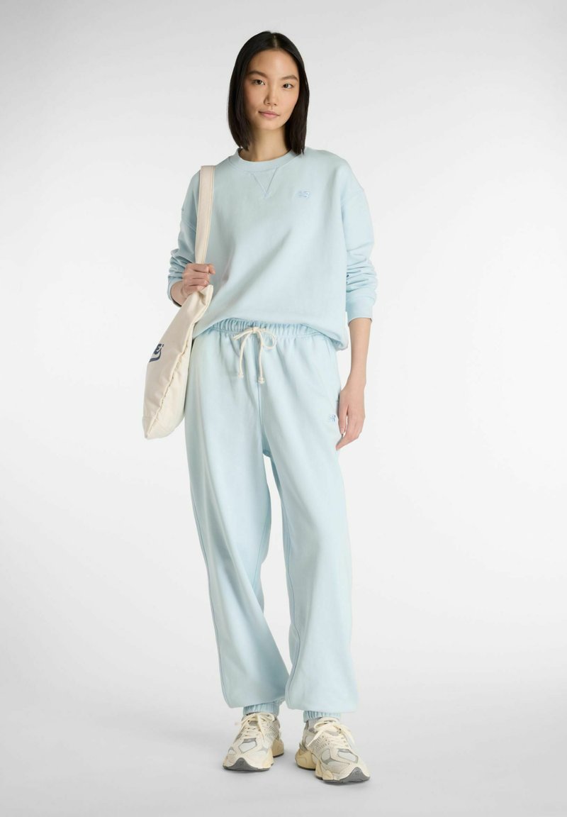 Felpa azzurro chiaro con scollo e polsini a coste, abbinata a pantaloni jogger coordinati con vita elasticizzata. Il modello tiene una borsa tote color crema.