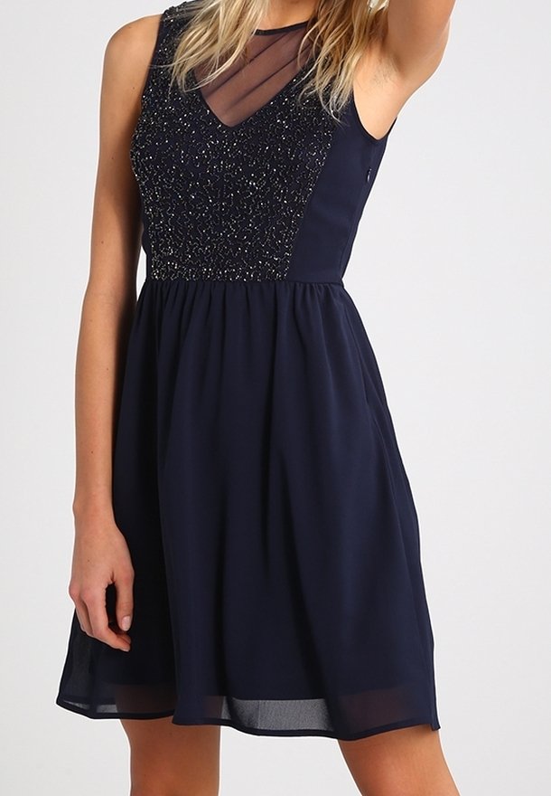 Robe sans manches bleu marine avec corsage perlé et panneau transparent en V, jupe fluide, portée par une personne aux cheveux blonds.