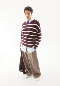 Weinrot und creme gestreifter Pullover über einem hellblauen Hemd mit Kragen, kombiniert mit weitem, braunem, strukturiertem Hosen; weiße Schuhe.