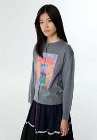 Cardigan gris avec un design graphique coloré sur le devant, fermetures à boutons et poignets côtelés. Porté par-dessus une jupe marine avec dentelle.