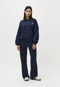 Donna in piedi che indossa una felpa con colletto a maniche lunghe blu navy e pantaloni coordinati, con sneakers bianche e nere su uno sfondo neutro.