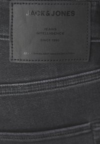 Jeans de denim negro con una etiqueta de cuero. La etiqueta presenta texto en relieve: "JACK & JONES", "JEANS INTELLIGENCE" y "CRAFTSMANSHIP AND DEDICATION."