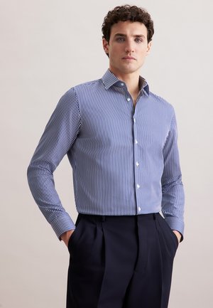 Mann in einem blau-weiß gestreiften Langarmhemd, das in hochgeschnittenen dunkelblauen Hosen steckt, mit Händen in den Taschen.