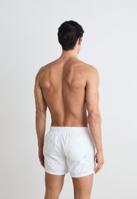 Shorts de bain blancs avec une taille élastique ; poche arrière avec une accentuation bleue ; modèle montré de dos, peau exposée.