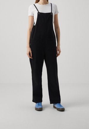 Schwarze Baumwolllatzhose mit verstellbaren Trägern, zwei großen Fronttaschen und lockerer Passform, kombiniert mit hellblauen Sneakers.