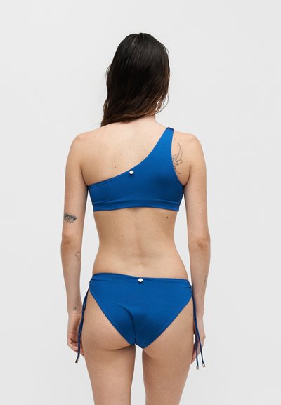 Γυναίκα με μακριά σκούρα μαλλιά, φοράει μπλε σουτιέν μαγιό με έναν ώμο και ταιριαστό κάτω μέρος με κορδονάκια, εμφανίζεται από πίσω με απλό φόντο.