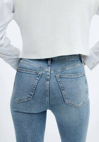 Femme portant un jean taille haute bleu clair et un pull court côtelé blanc, les mains reposant sur les hanches, vue de dos.