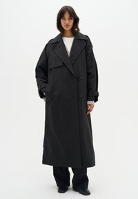 Manteau noir en trench avec un design à double boutonnage, de larges revers et des poches latérales. Le tissu semble lisse et légèrement brillant.
