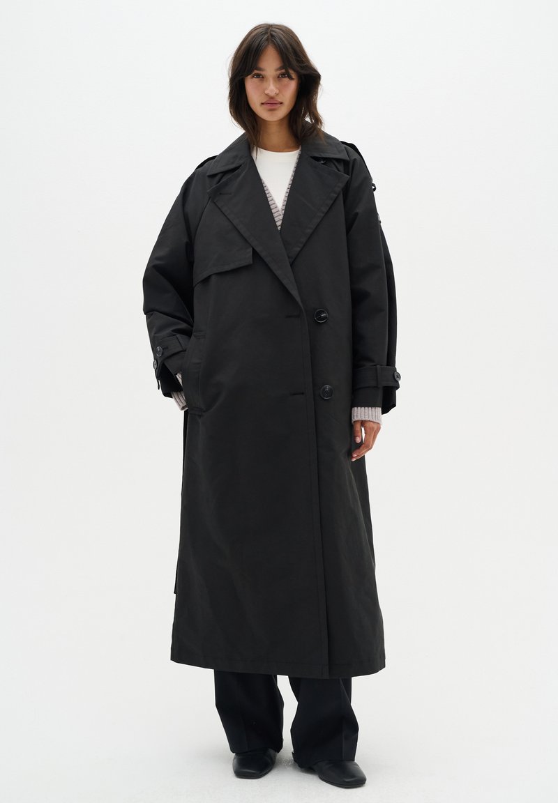Manteau noir en trench avec un design à double boutonnage, de larges revers et des poches latérales. Le tissu semble lisse et légèrement brillant.