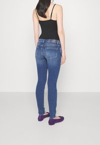 Vaqueros ajustados azul con muslos deslavados, una cinturilla oscura y dos bolsillos traseros. Combinados con zapatos planos morados y una camiseta sin mangas negra.