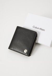 Black leather wallet with a zippered closure and silver hardware, featuring a round logo accent and accompanied by a white box with branding.